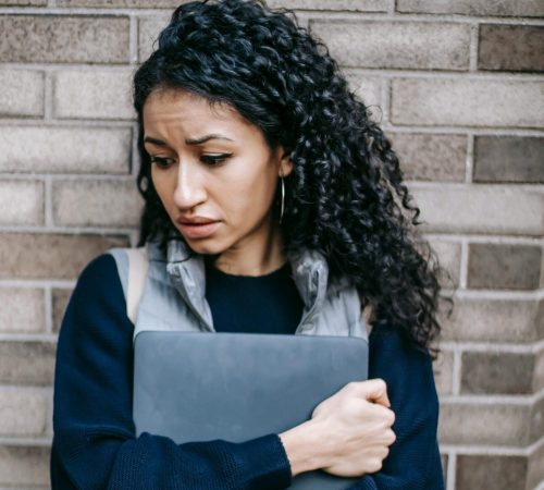 Tired Hispanic female with curly hair leaning on brick wall while carrying netbook and looking down